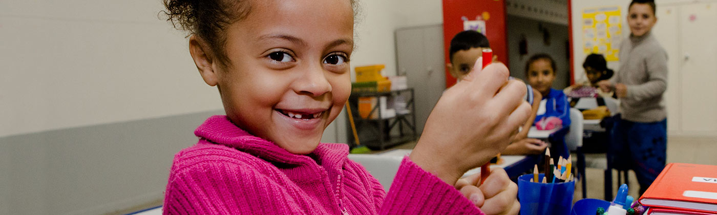 Criança, segurando um lápis e sorrindo para a câmera, dentro de uma sala de aula e com colegas ao fundo