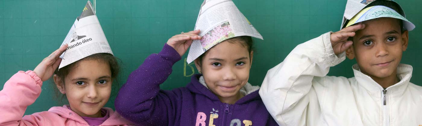 Três crianças em uma sala de aula em frente a uma lousa, com chapéus de papel e fazendo o símbolo da continência
