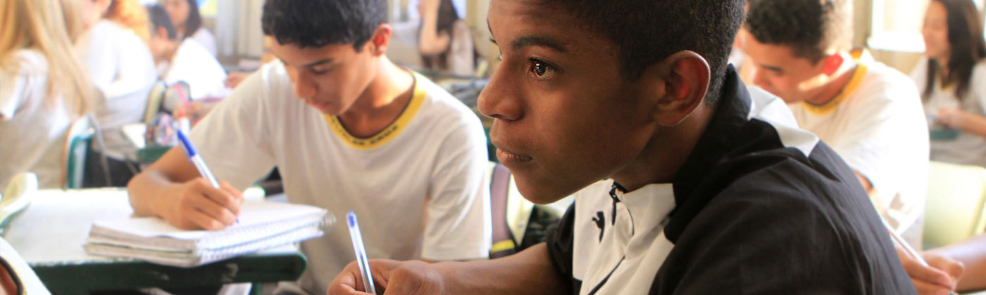 Alunos estudando dentro de uma sala de aula com foco em um garoto anotando no caderno e olhando para frente