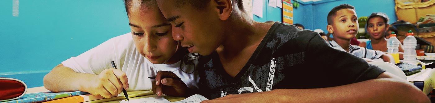 Imagem mostra duas crianças (um menino e uma menina) sentados numa sala de aula tentando resolver um exercício no caderno