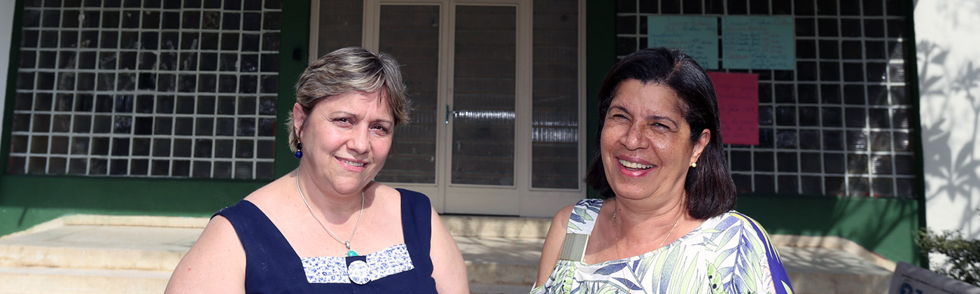 Duas mulheres no pátido de uma escola, sorriem para a foto