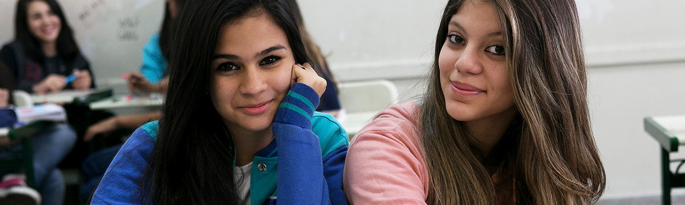Imagem mostra duas alunas dentro de uma sala de aula, com foco nos rostos e sorrindo para a foto